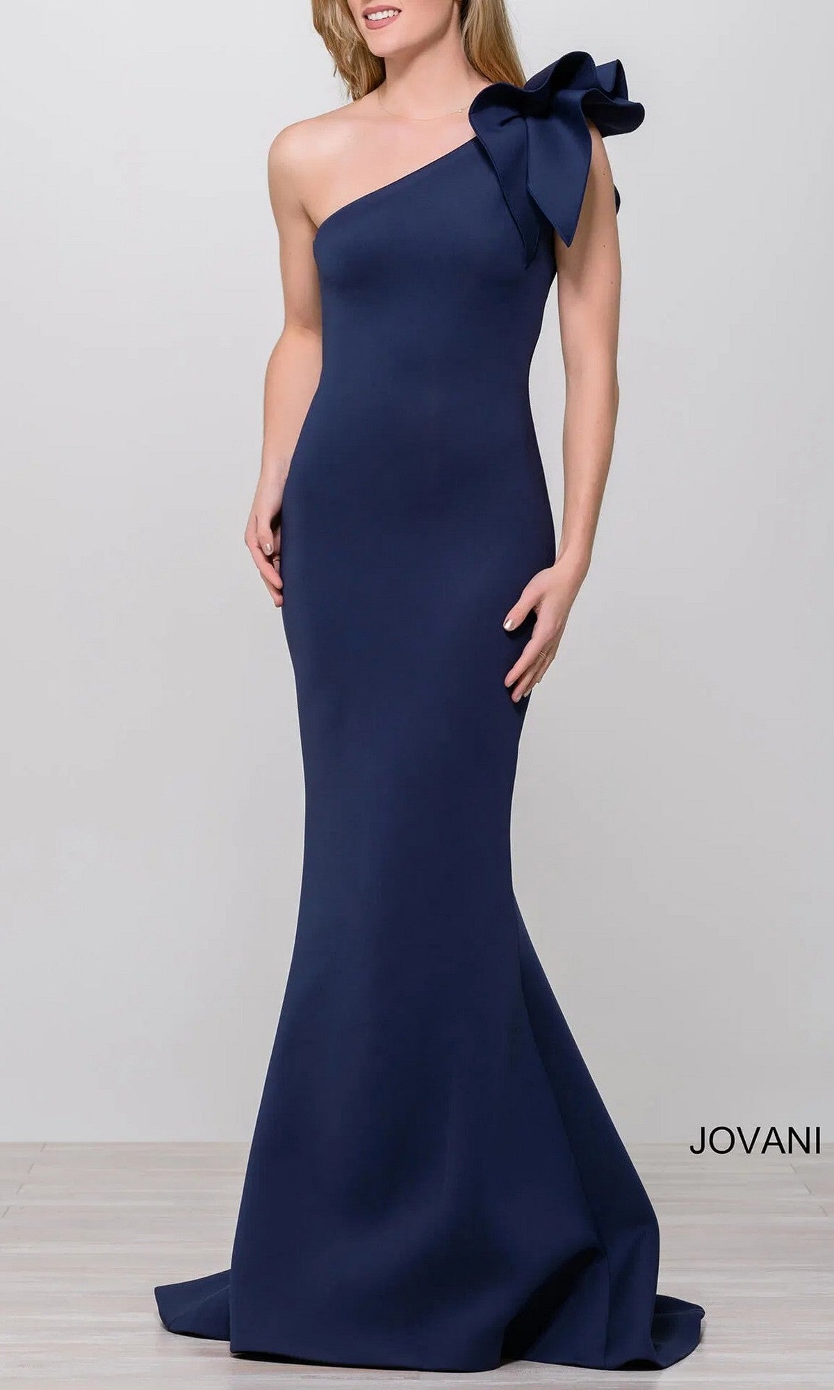 A woman models the Jovani 32602 One-Shoulder Mermaid Prom Gown in navy blue, featuring a dramatic shoulder bow; “Jovani” is displayed in the bottom right corner.